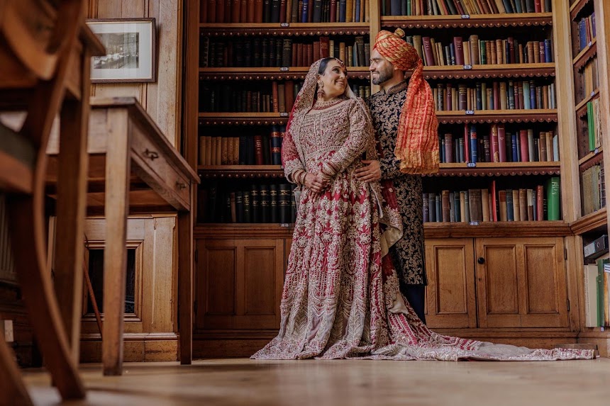 A couple in traditional attire stands in a warmly lit room with wooden bookshelves. The woman wears an ornate dress, and the man, in a complementary outfit with a turban, holds her from behind. They gaze lovingly at each other in Dulwich old libray